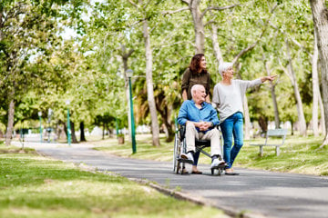 A caregiver and her wheelchair-using patient walk in a park with the patient's wife as additional support.