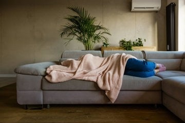 Woman hiding face under blanket on sofa struggling with migraine postdrome