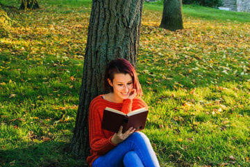 Young woman sitting under a tree reading stories to help her escape cancer trauma