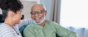 A smiling black couple sits together on a couch, embodying joy and support in the context of caregiving.