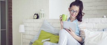 A woman sits on a bed, holding a tablet and a cup of coffee, embodying self-kindness amidst anxiety.